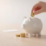 Weiße Spardose in Schweinchenform, in die eine Hand eine goldene Münze einwirft, daneben gestapelte Münzen auf einem Holztisch.