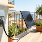 Ein Schuko-Stecker steckt in einer Außensteckdose auf einem sonnigen Balkon vor einer Mini-Solaranlage.